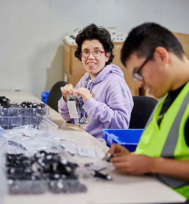 A woman in a lilac hoodie packing small parts at a long workbench, a colleague in a hi-vis vest working beside her