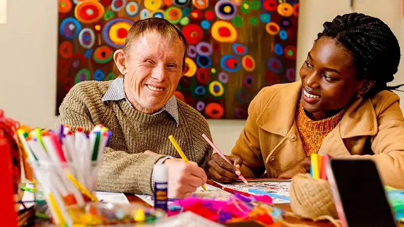 Two people smiling at a table covered in pens and art supplies